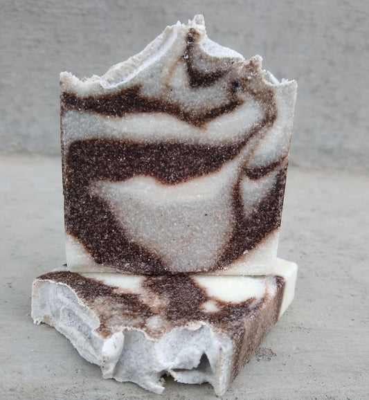 An image of one bar of Backyard Quarry soap sitting on top of a second bar of soap. There is a plain background that contrasts with the swirls of gray and white of the soap